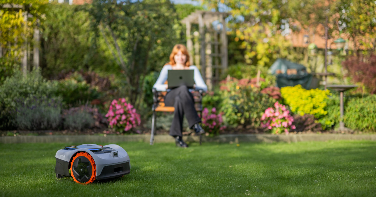 Robotfűnyíró nyírja a füvet egy gondozott kertben, a háttérben egy nő ül, laptopján dolgoz